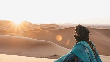 mujer con sudadera azul sentada en la arena durante la puesta de sol